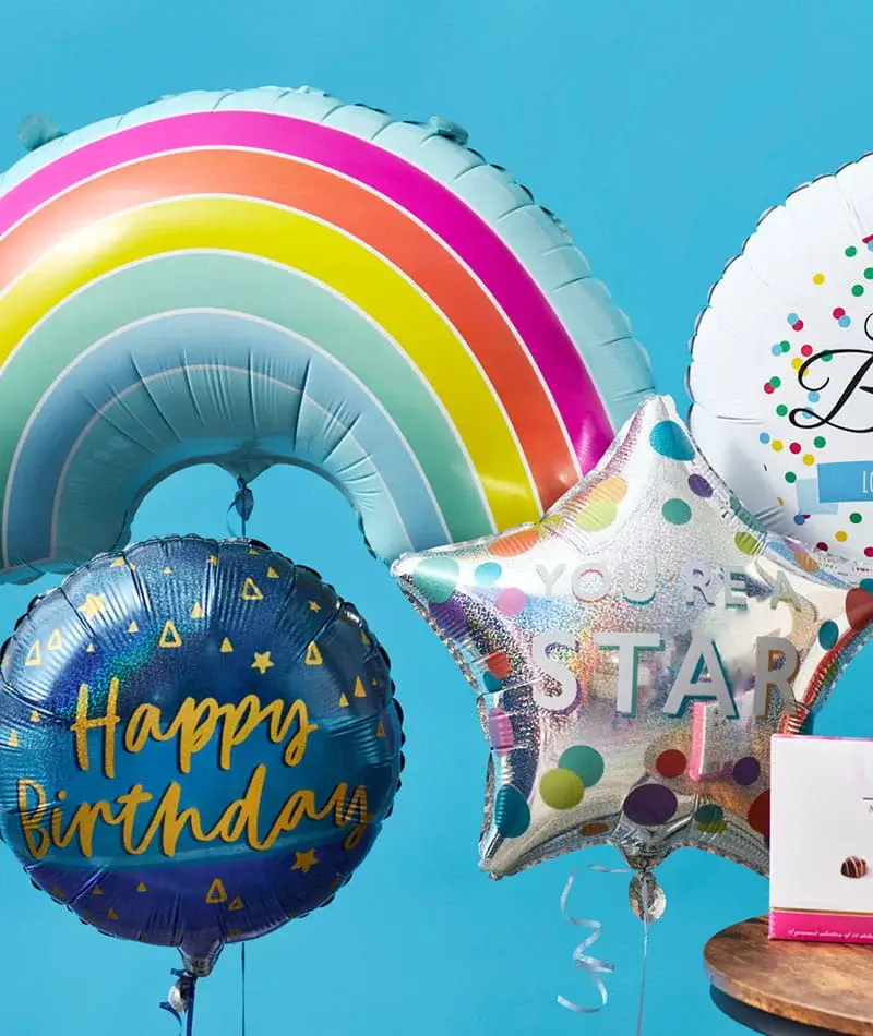 A colourful arrangement of foil balloons including a rainbow, a blue Happy Birthday balloon, a silver star balloon with polka dots and a white balloon with confetti-style details, all set against a bright blue background.